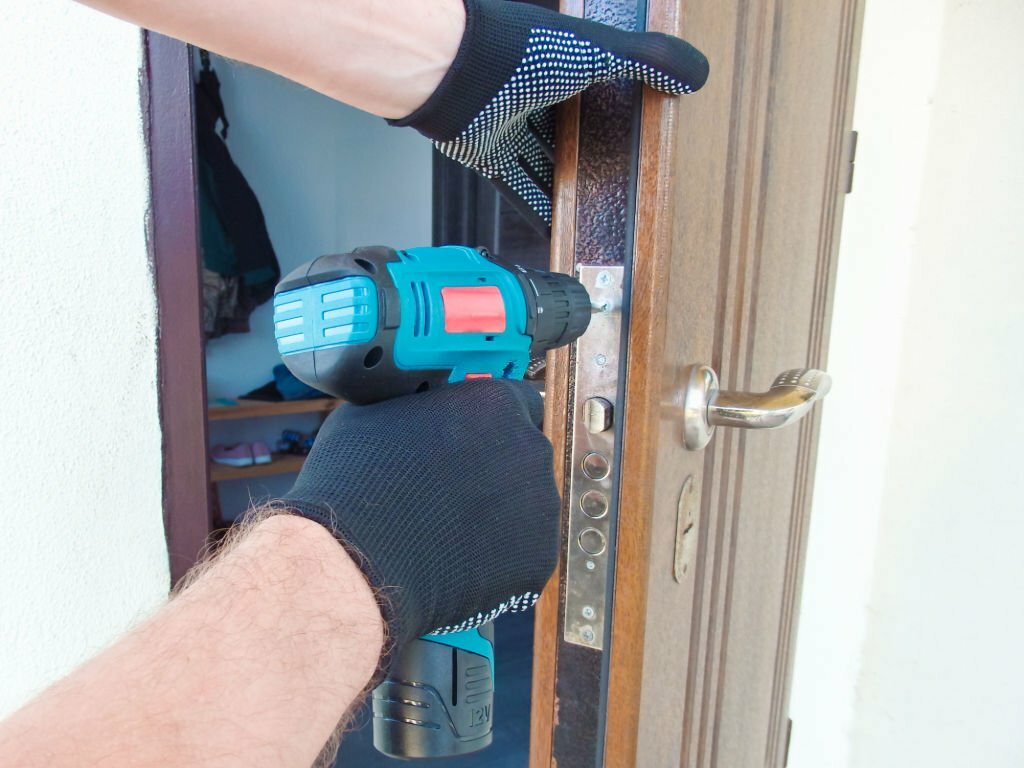 The master worker installs a door lock in the front door, metal doors with a polymer coating, using an electric drill screwdriver, close-up.