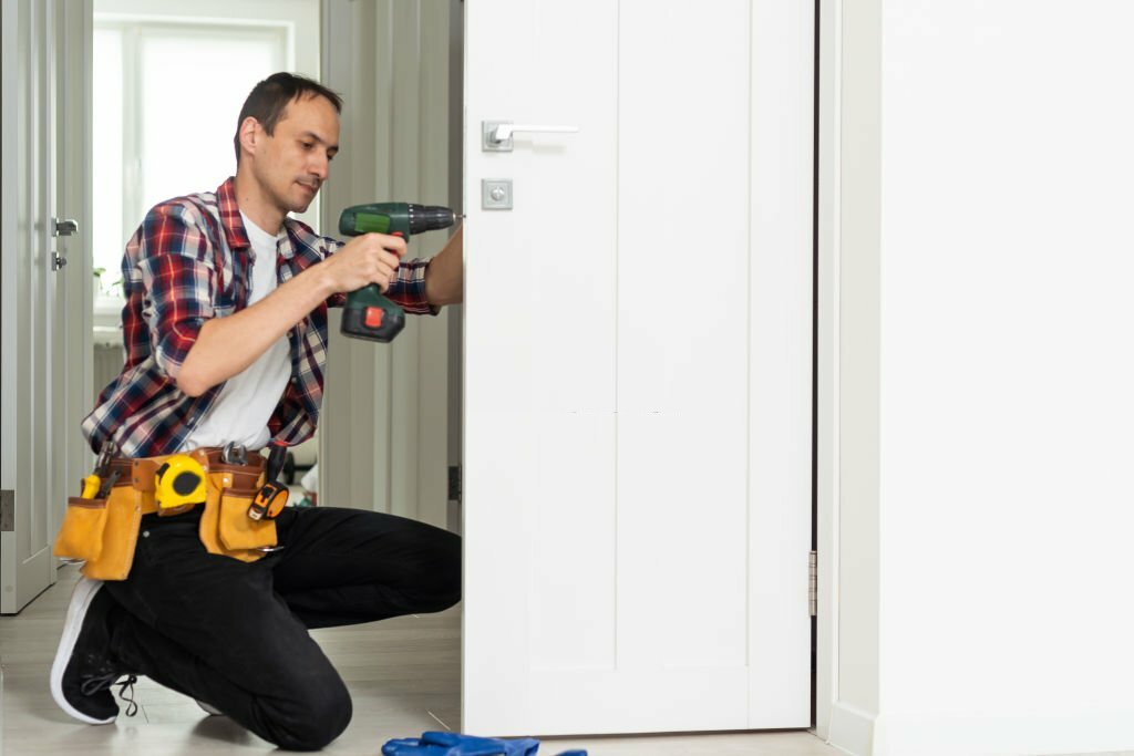 Maintenance Concept. Focused serious young locksmith with tattoo on hand wearing blue uniform standing on one knee in doorway installing doorknob handle, repairing wooden front door entrance. High quality photo