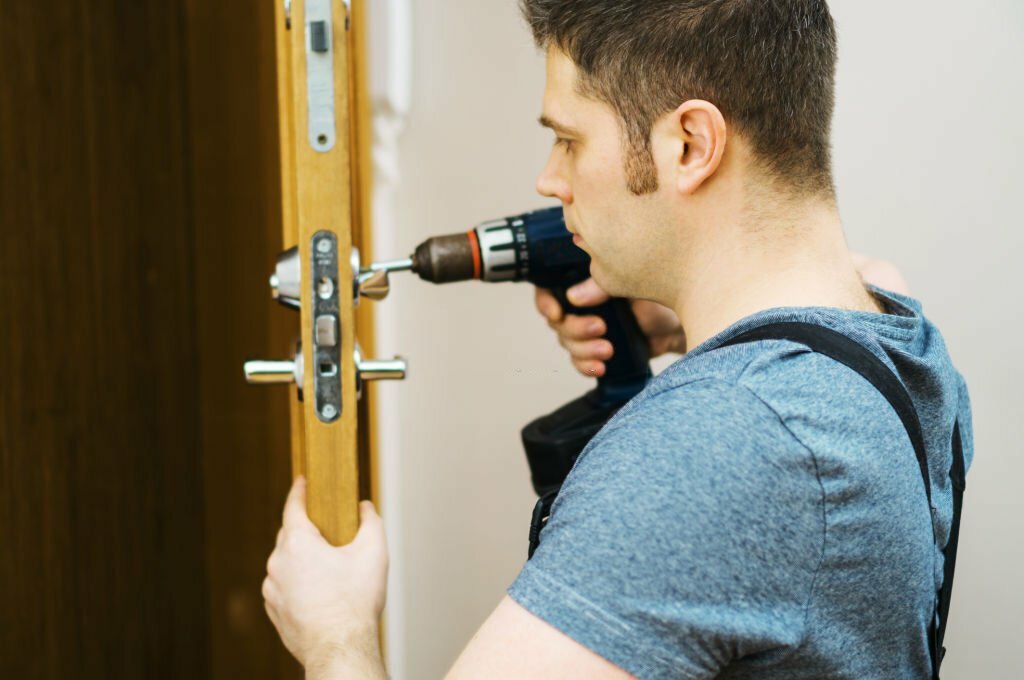 Young handyman in uniform changing door lock.