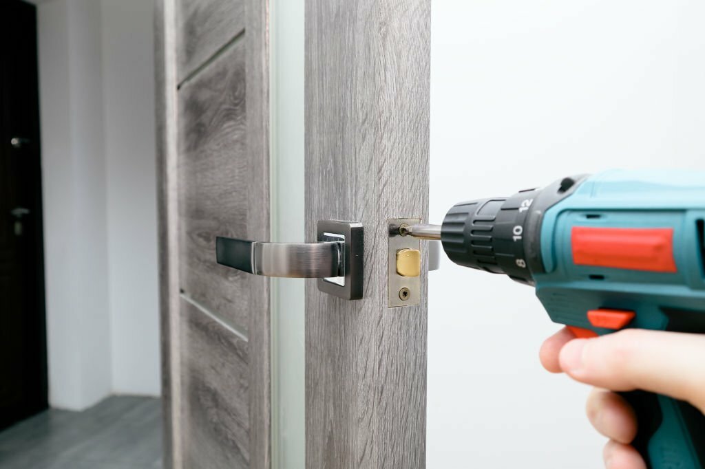 A man repairing a doorknob using a screwdriver at home. Carpenter at lock installation with electric drill into interior wood door