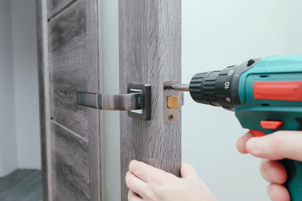 A man repairing a doorknob using a screwdriver at home. Carpenter at lock installation with electric drill into interior wood door